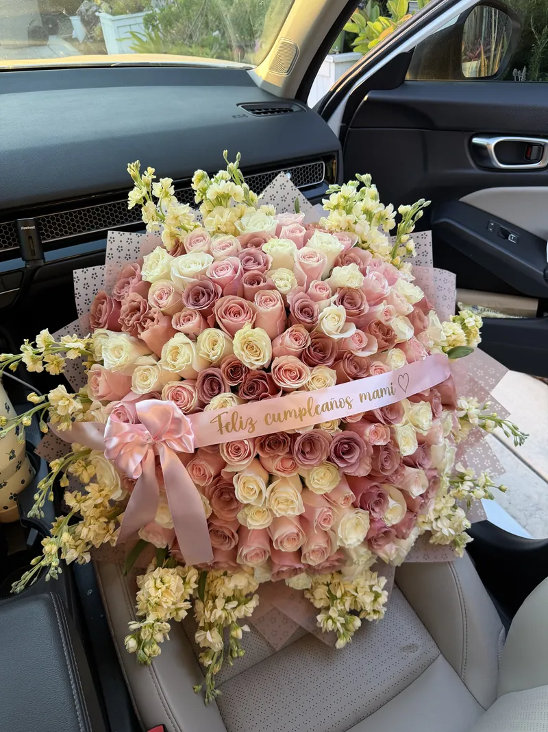 A large blush and cream spray-rose bouquet with yellow stock and a satin sash reading Feliz cumpleaños mami.
