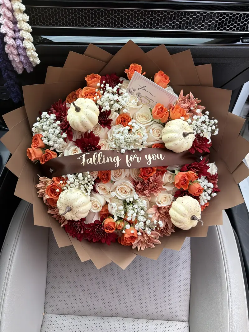 Cream and orange roses with burgundy chrysanthemums, baby&rsquo;s breath and small white pumpkins in a kraft paper wrap, tied with a brown satin sash reading Falling for you.