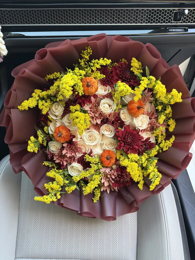 Cream roses with burgundy and dusty-rose chrysanthemums, sprays of bright yellow goldenrod and small orange pumpkins, in a deep burgundy ruffled wrap.