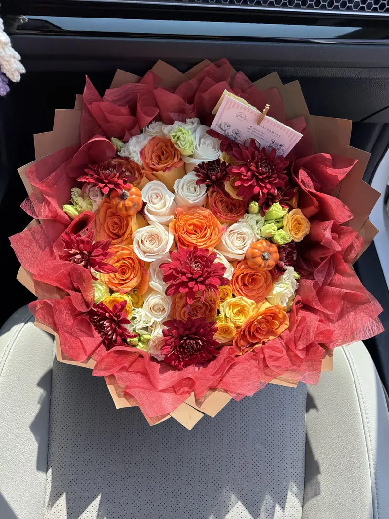 A round autumn bouquet of orange, cream and yellow roses with deep burgundy chrysanthemums and two small orange pumpkins, wrapped in red tissue over kraft paper.