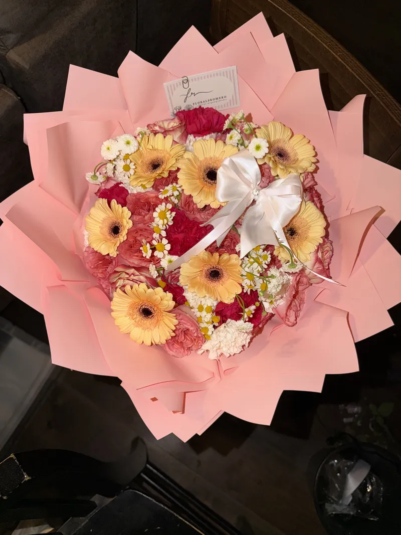 Yellow gerbera daisies, carnations and chamomile in a petal-folded blush wrap with a white satin bow.