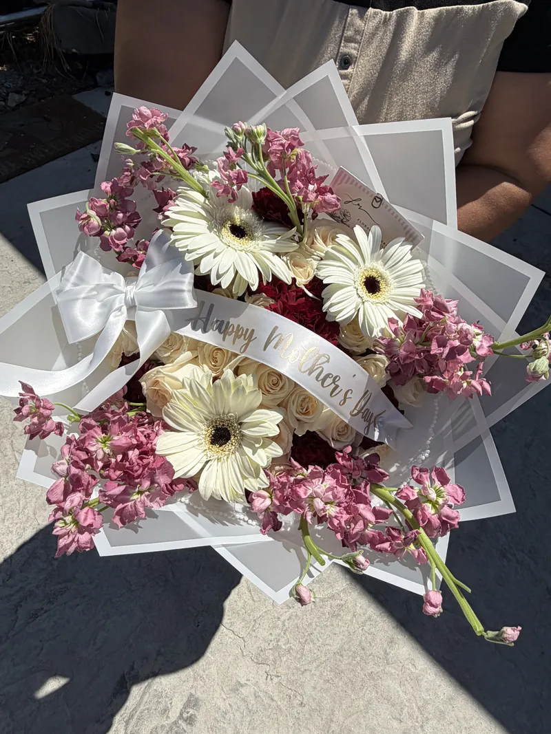 White gerbera daisies, cream roses and pink stock in a frosted wrap with a Happy Mother's Day sash.