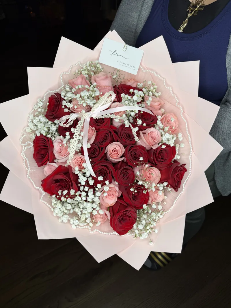 A round bouquet of deep red and soft pink roses with baby's breath, wrapped in blush paper and edged with pearl trim.